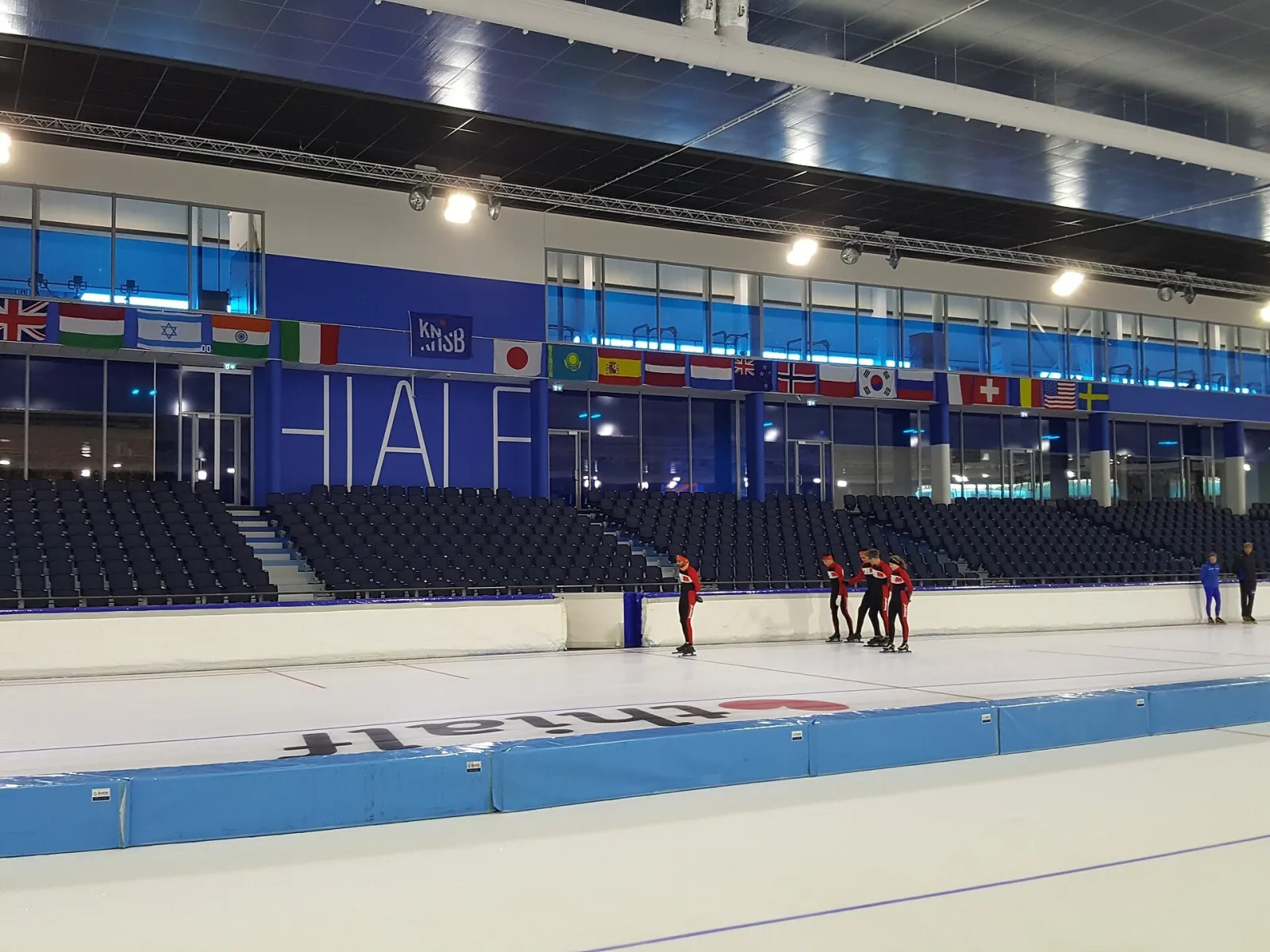 Schaatsers op het ijs in het Thialf-stadion, met erachter de blauwe tribunes, de vlaggen en de glazen bovenverdiepingen van de hal. – Thialf