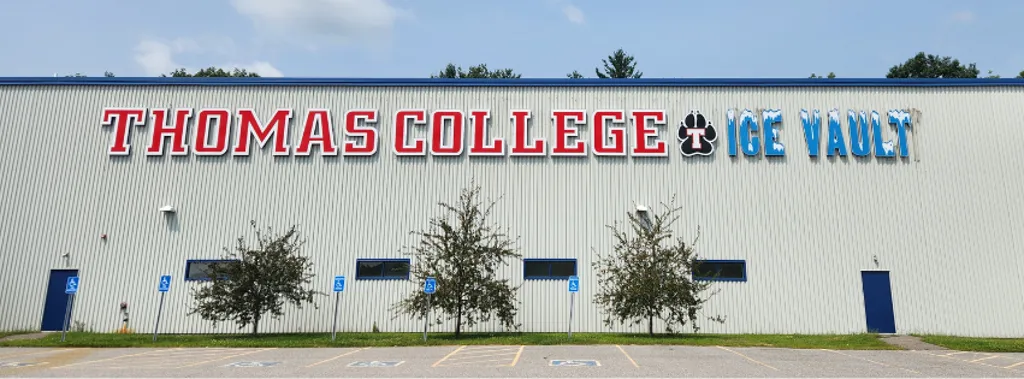 Front of the metal-sided building with a large "THOMAS COLLEGE ICE VAULT" sign along the roofline, accessible entrances and parking below. — Thomas College Ice Vault