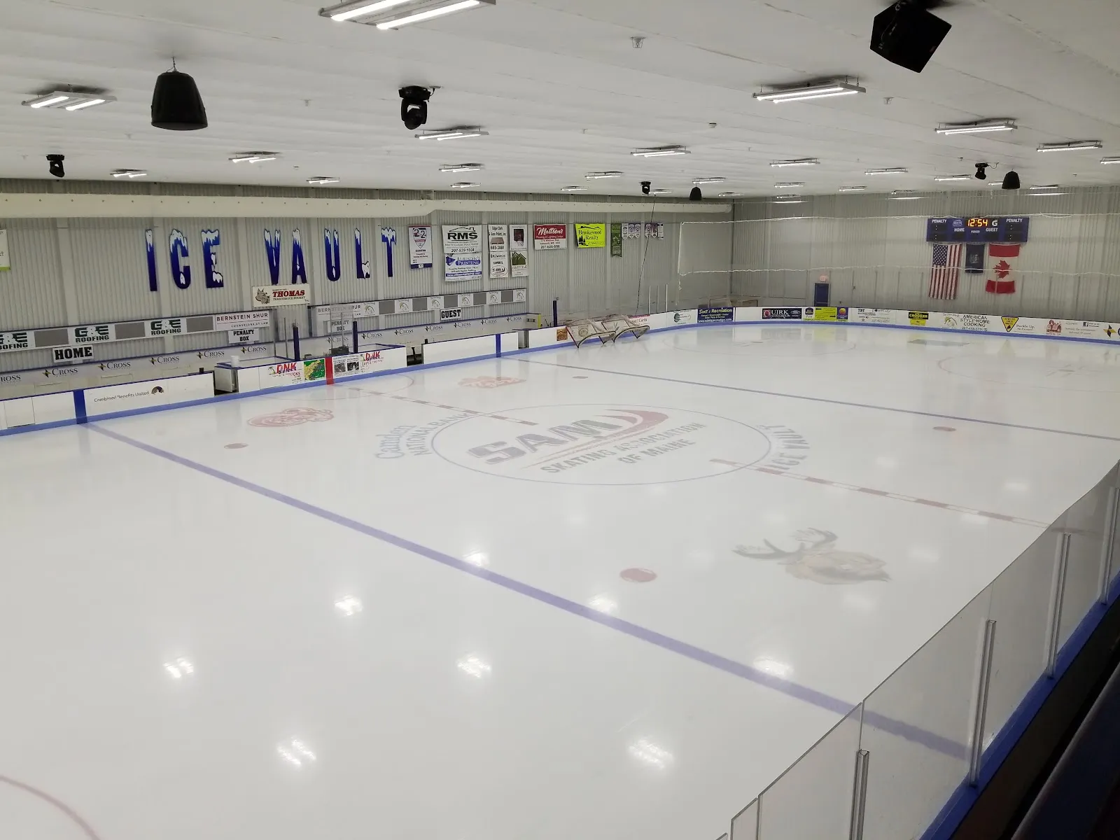 Empty ice sheet from the stands, with "ICE VAULT" lettered on the end wall, sponsor boards, a scoreboard, and the SAM (Skating Association of Maine) logo painted at center ice. — Thomas College Ice Vault