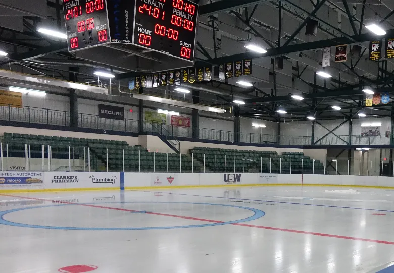 The empty ice surface of the Thompson Regional Community Centre arena, with the scoreboard above the boards and the stands along the far side. — Thompson Regional Community Centre