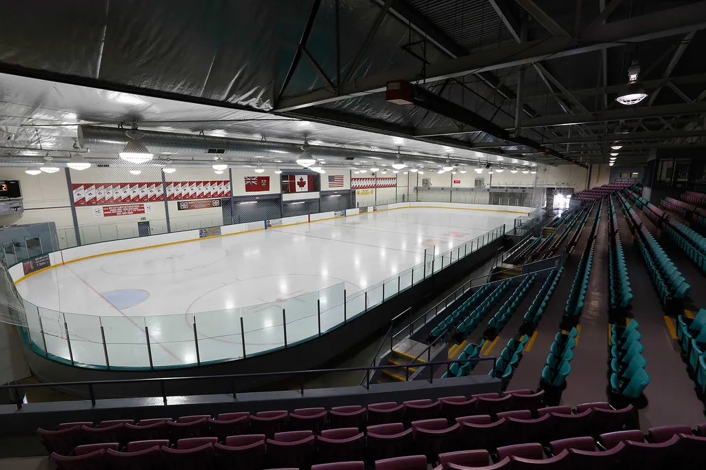 Full view of an ice sheet at Thornhill Community Centre's arena from the upper seating area, showing empty teal and burgundy stadium seats, the boards and glass, and Canadian/Ontario/US flags on the far wall. — Thornhill Community Centre