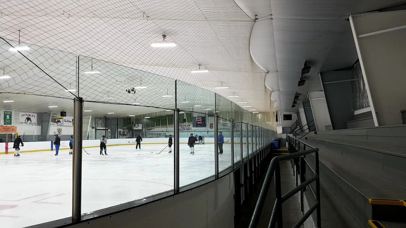 Una vista amplia a lo largo de un barandal oscuro de la pista, con un partido de amarillo y blanco a lo lejos sobre el hielo bajo un techo claro arqueado. — Thornton Community Centre (Essa)