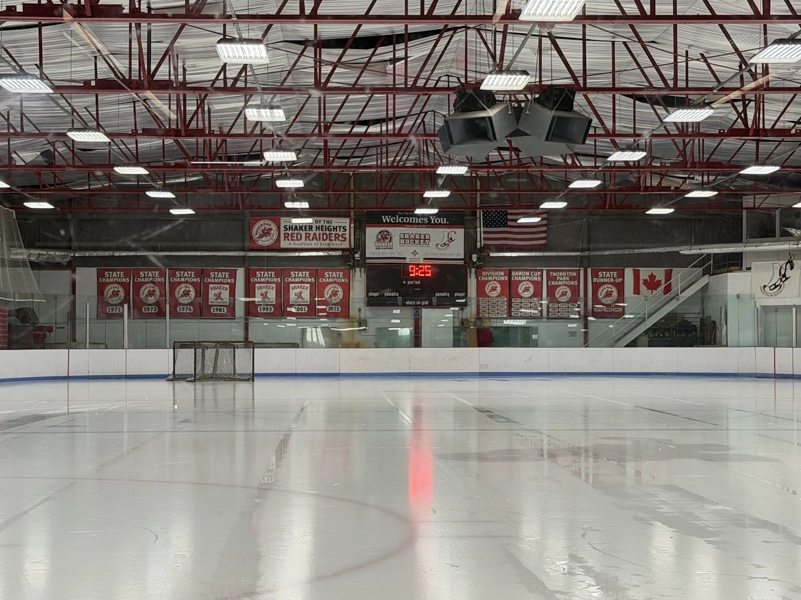 Una pista vacía con una línea central roja y un círculo de saque bajo un techo de vigas de acero rojas, con una hilera de banderines colgados en la pared del fondo. — Thornton Park Ice Arena