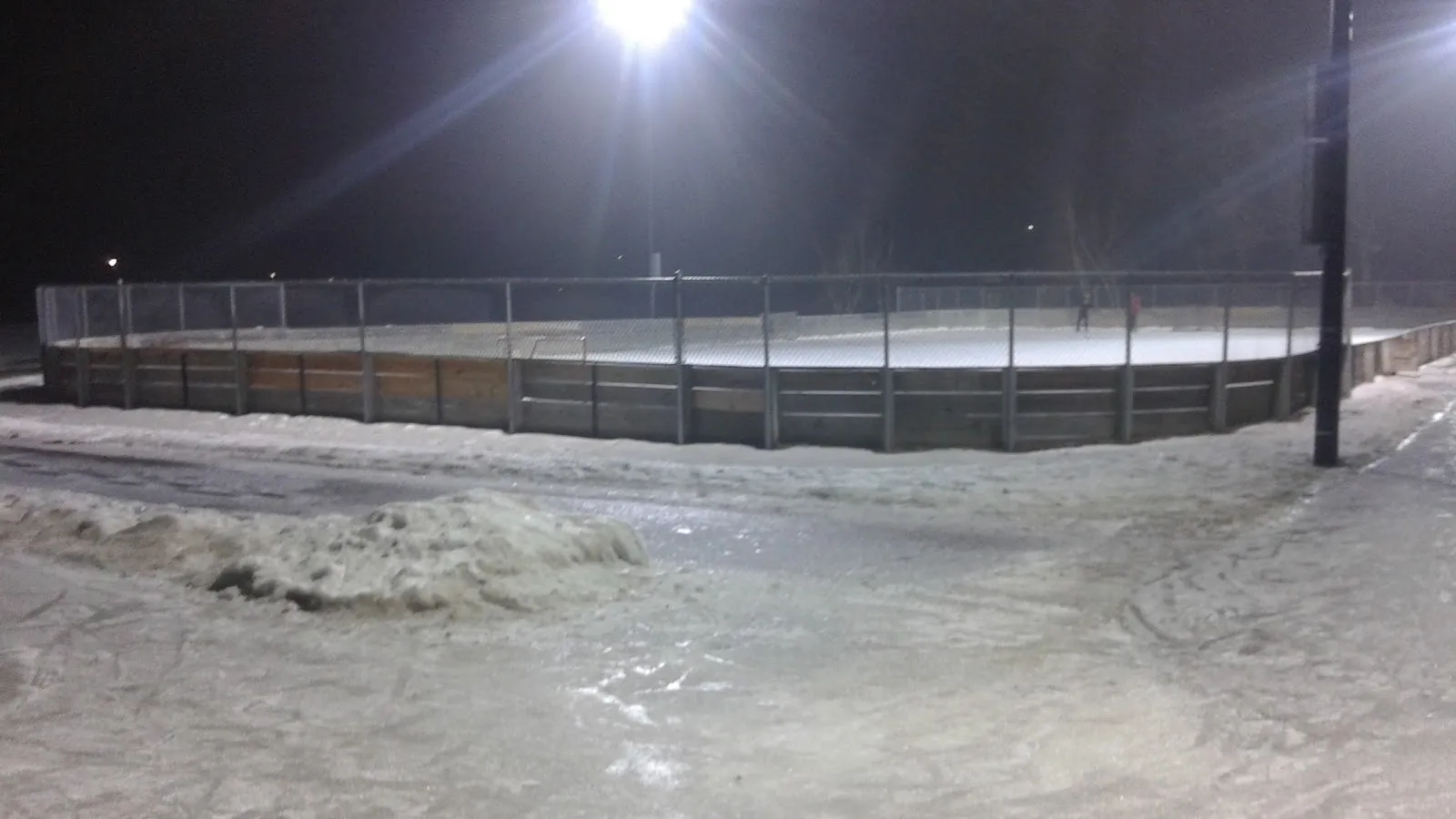 The boarded outdoor rink at Thorpe Park lit by floodlights at night, seen across a snowy path, with wooden boards, fencing and ice on the sheet; no people. — Thorpe Park Ice Rinks
