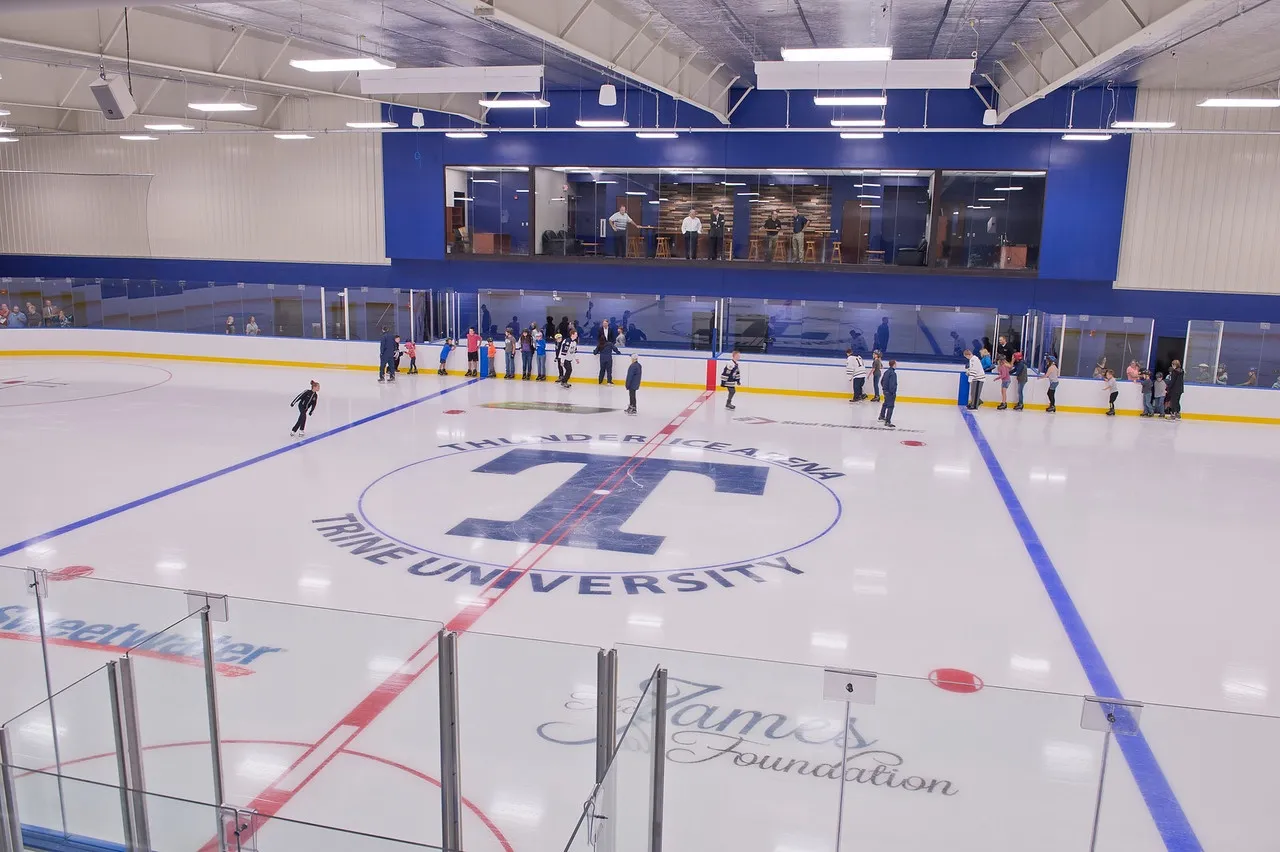 Una vista amplia del hielo con la T de la universidad en el centro, unos cuantos patinadores junto al vidrio y una galería de prensa azul y banderines en la pared del fondo. — Thunder Ice Arena (Angola)