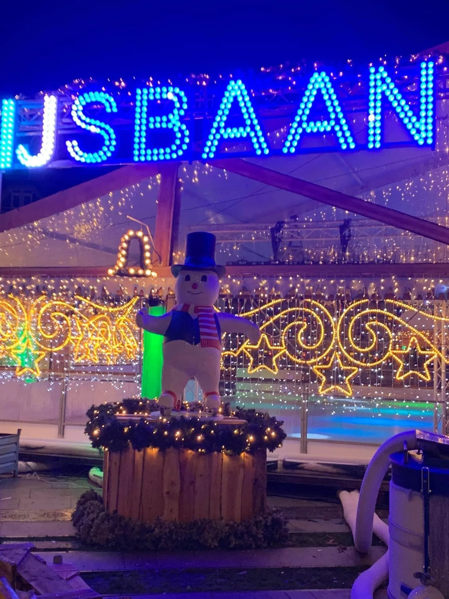 The illuminated IJSBAAN sign and a decorated snowman at the Tilburgs Winterparadijs rink, lit up at night under the tent roof — Tilburgs Winterparadijs