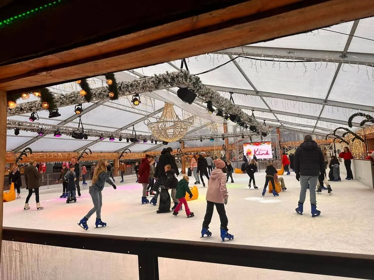 Skaters of all ages spread across the white ice of the tent-covered rink of the Tilburgs Winterparadijs, seen from the rail with the lit tent roof above — Tilburgs Winterparadijs