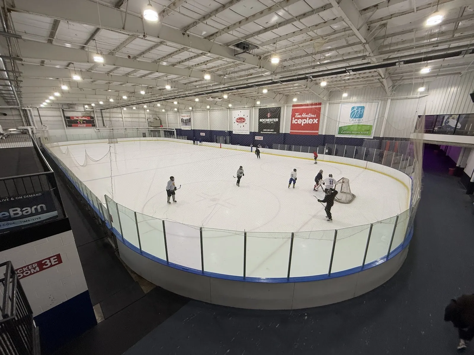 A wide fisheye view of a sheet curved round the boards, skaters spread across the ice under a steel-trussed roof with sponsor signs along the far wall. — Tim Hortons Iceplex