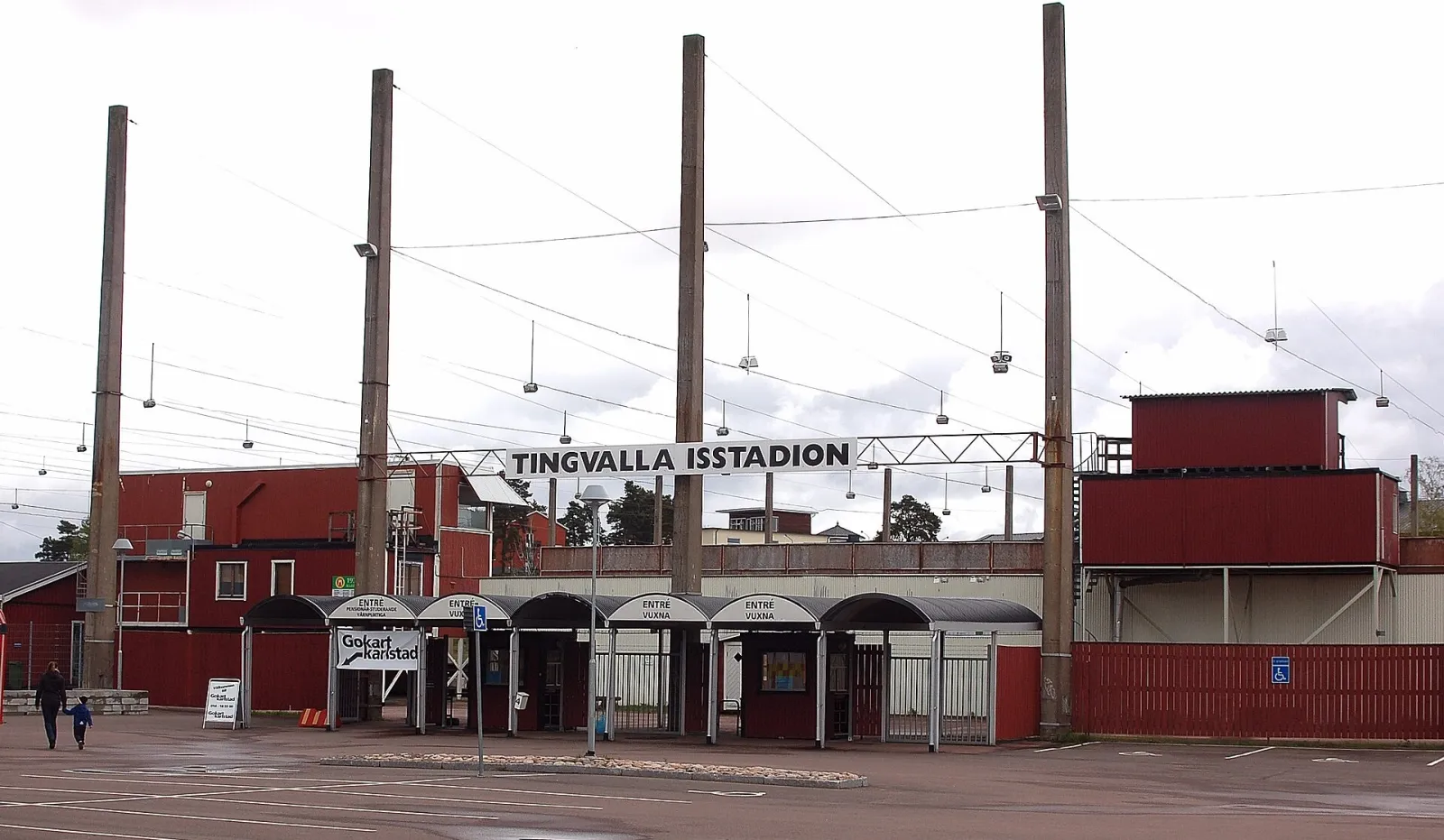 The red entrance gate of Tingvalla Isstadion with its name on the sign above the arches and the tall floodlight masts behind. — Tingvalla isstadion