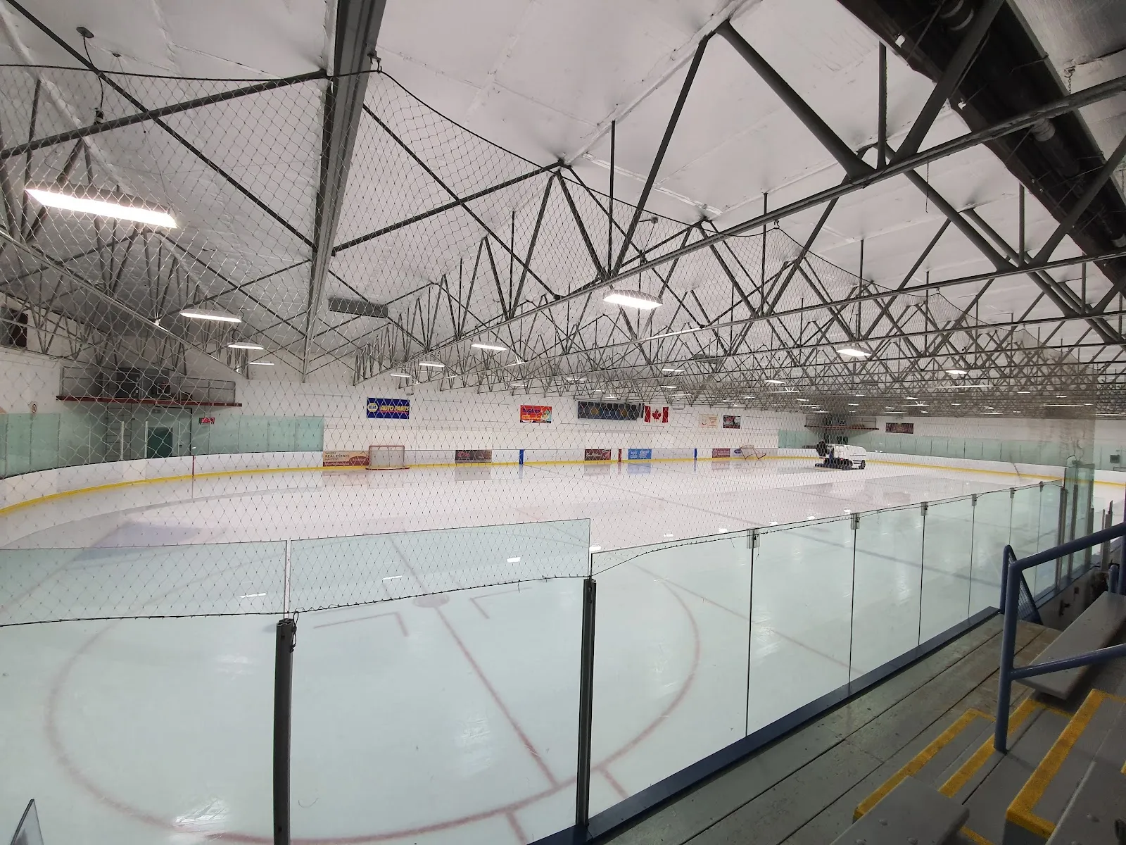 Empty indoor ice rink viewed from the stands, full length of the ice sheet visible with a Zamboni parked at the far end and sponsor boards along the boards, no people present. — Tipton Arena