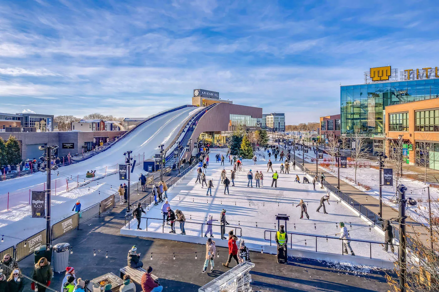 Skaters on the Hy-Vee Plaza rink in daylight, with the Ariens Hill tubing ramp rising behind the ice and the Titletown buildings along the plaza. — Titletown Ice Rink at Hy-Vee Plaza