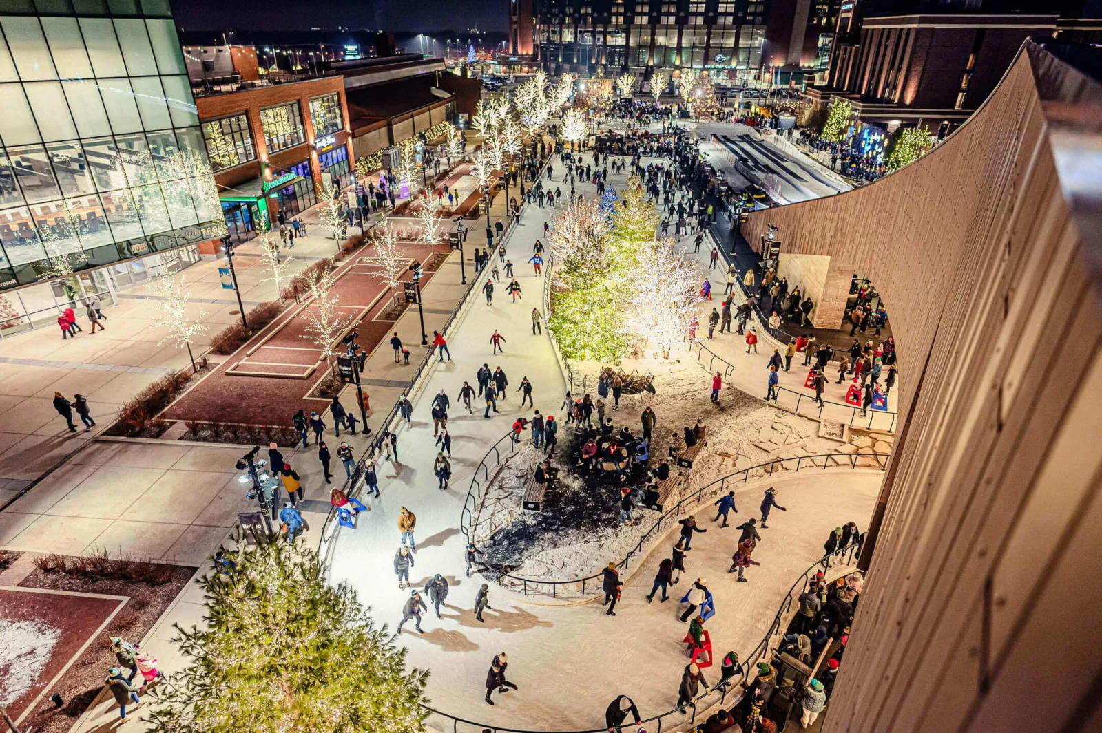 The Hy-Vee Plaza rink at night from above, with skaters circling the ice trail around the fire-pit island between the plaza buildings. — Titletown Ice Rink at Hy-Vee Plaza