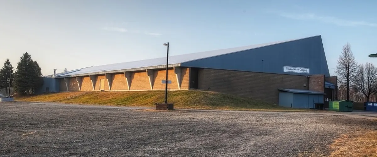 The long brick arena with its sloped blue roof and entrance, across a gravel lot. — T.M. Davies Community Centre and Arena (Lively)