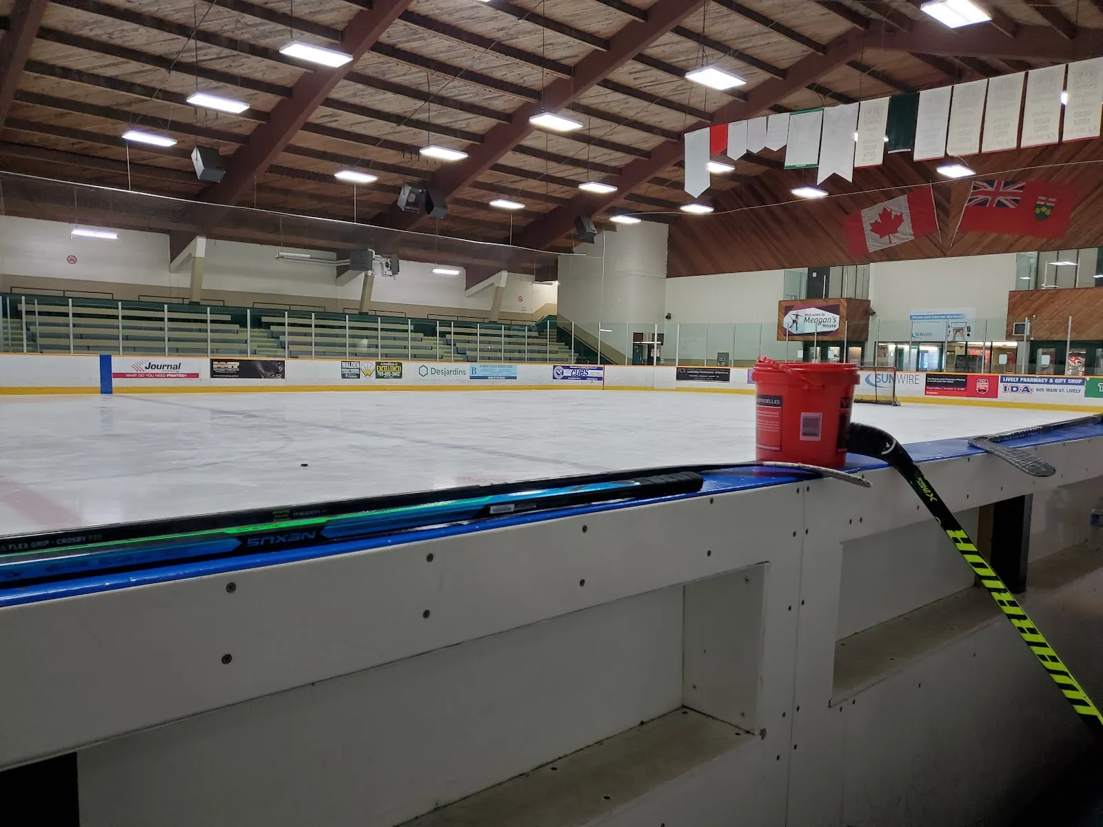 The ice seen over the bench boards, under the wooden roof beams with flags and banners on the far wall. — T.M. Davies Community Centre and Arena (Lively)
