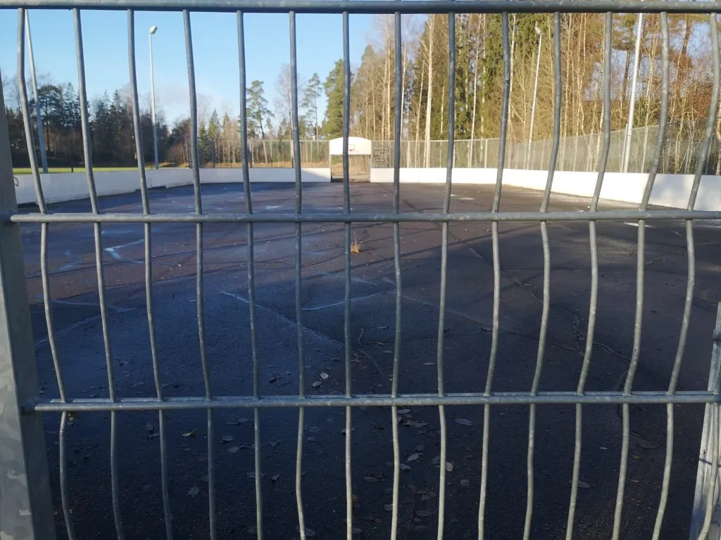 The Toija hockey rink seen through its steel fence in autumn, a bare paved surface with trees beyond. — Toijan kaukalo ja luistelukenttä (Salo)