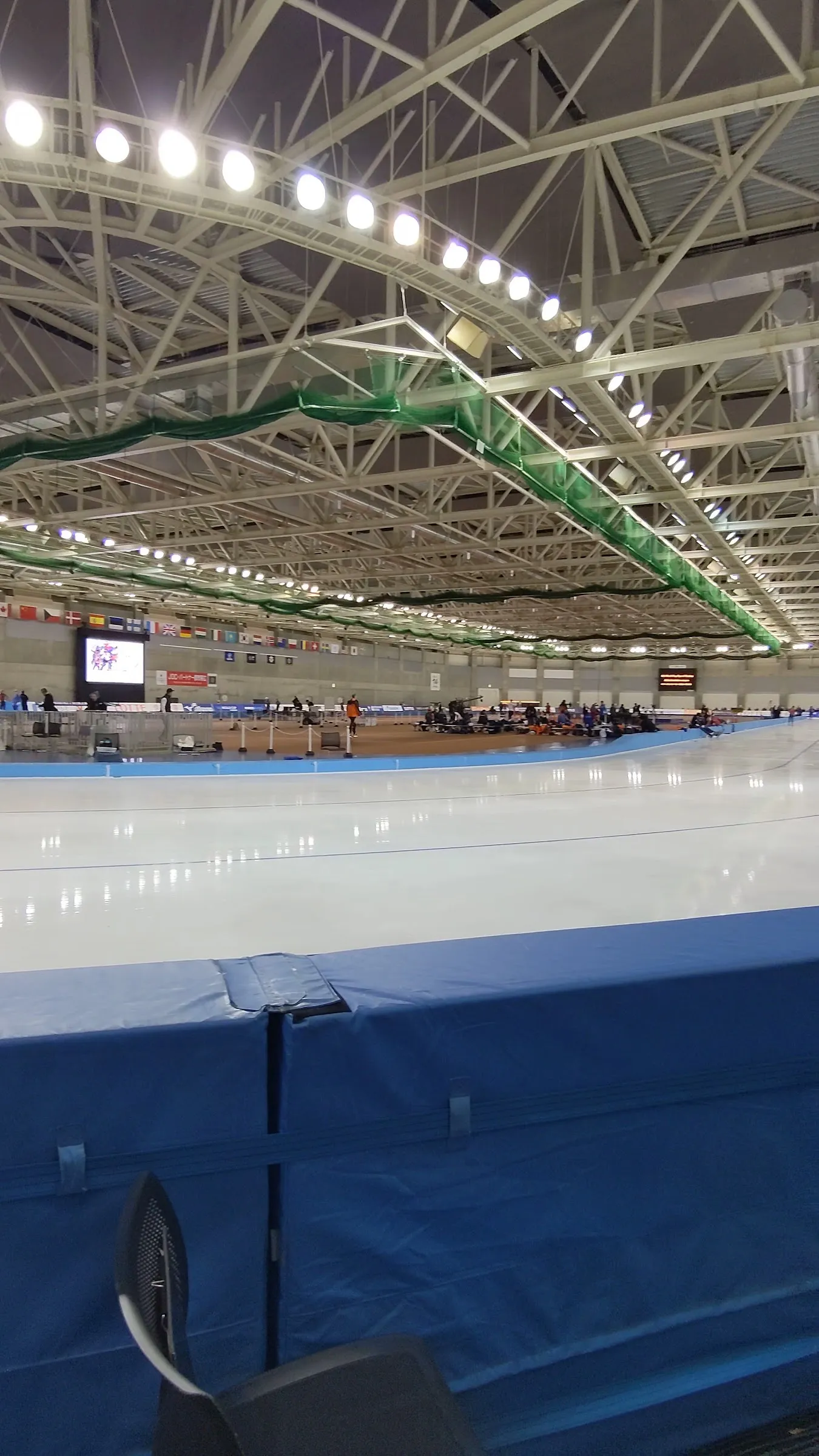 フェンス越しに見た十勝オーバルの400mの氷面。頭上には投光器の輪と鉄骨のトラス屋根が見えます。 — Tokachi Oval