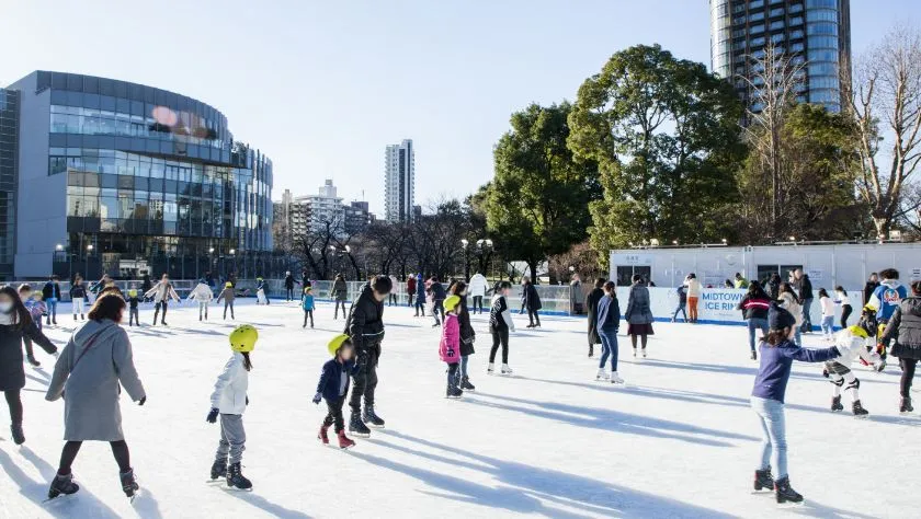 The open-air Midtown ice rink on the lawn in daylight in the 2025-26 season, with skaters spread across the ice and the glass Suntory museum building behind. — Tokyo Midtown Ice Rink