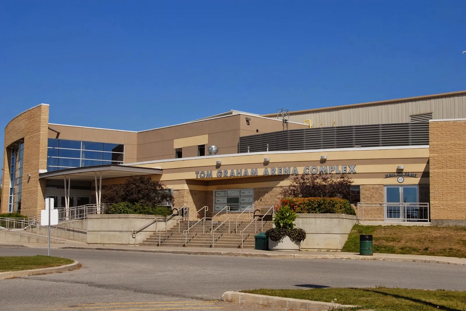 The tan-brick Tom Graham Arena Complex entrance under a clear sky, with the name over the doors. — Tom Graham Arena Complex (Richmond Hill)