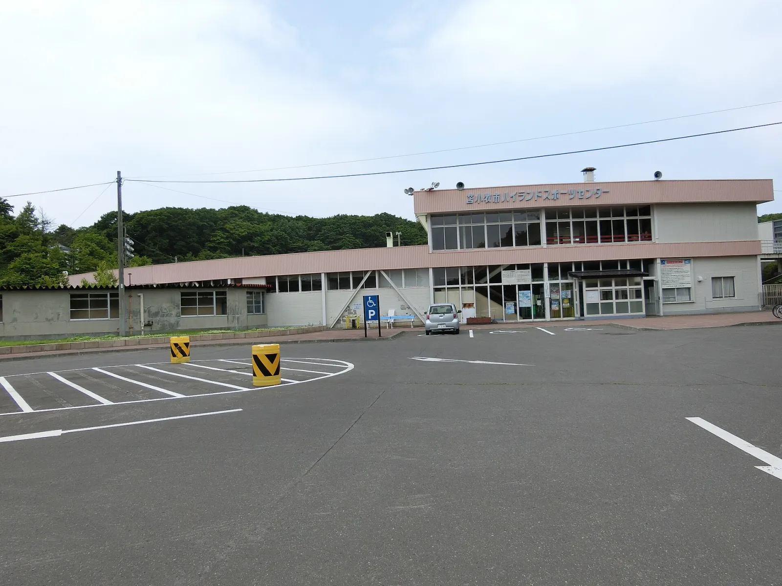 駐車場越しに見える2階建てのハイランドスポーツセンター。入口脇には身障者用駐車スペースがあり、背後には木々の茂る丘が広がっています。 — Tomakomai Highland Sports Center (Speed Skating Oval)
