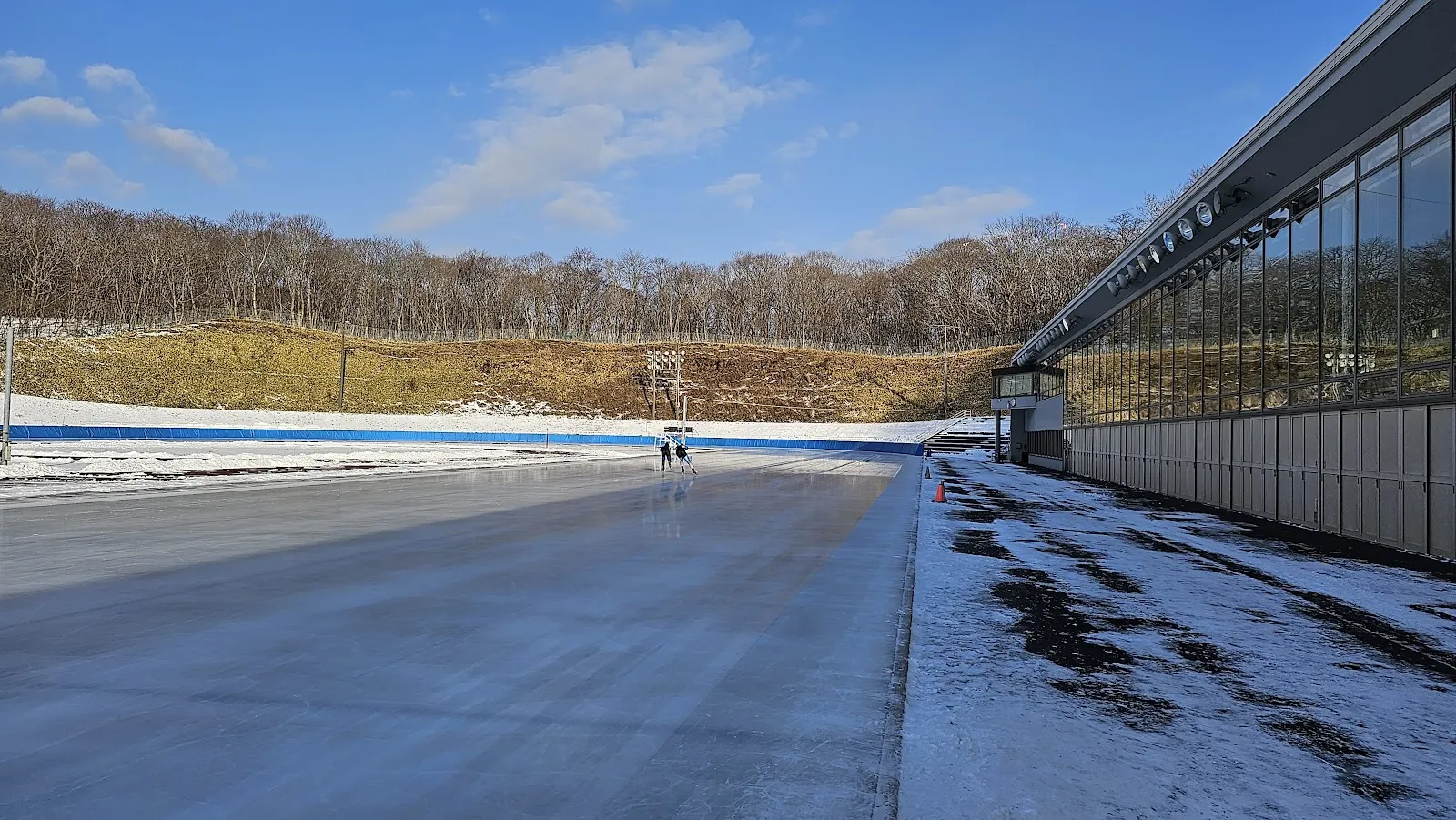 ガラス張りのスタンド棟に沿ったホームストレートから見た400mスピードスケート場。縁には雪が残り、背後の丘は葉を落としています。 — Tomakomai Highland Sports Center (Speed Skating Oval)