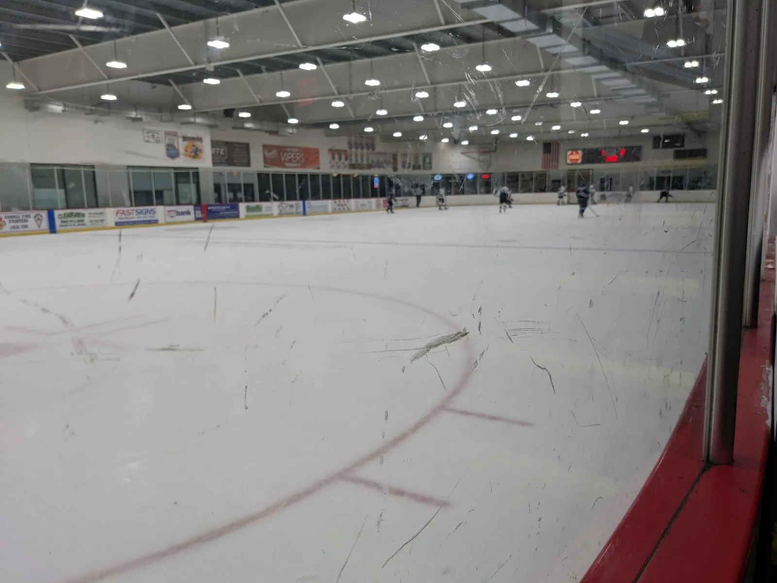 A wide view of the sheet through the glass from the corridor, the white ice and red lines with a few players far down the rink. — Top Shelf Ice Arena