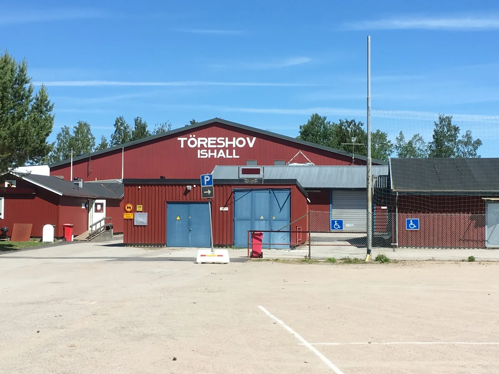 Den röda trähallen Töreshov ishall i Töreboda med namnet på gaveln, blå sidodörrar och en tom grusplan framför under blå himmel – Töreshov ishall