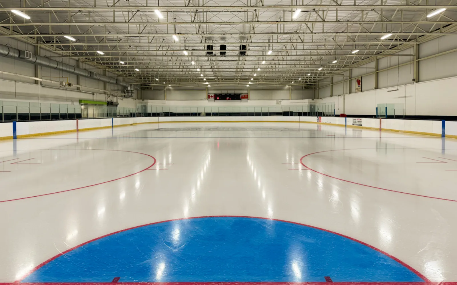 Toda la pista vista a nivel del hielo desde un extremo, con el área de portería azul y las marcas rojas del centro de la pista, y el marcador y una bandera de Canadá visibles al fondo. — Albion Arena