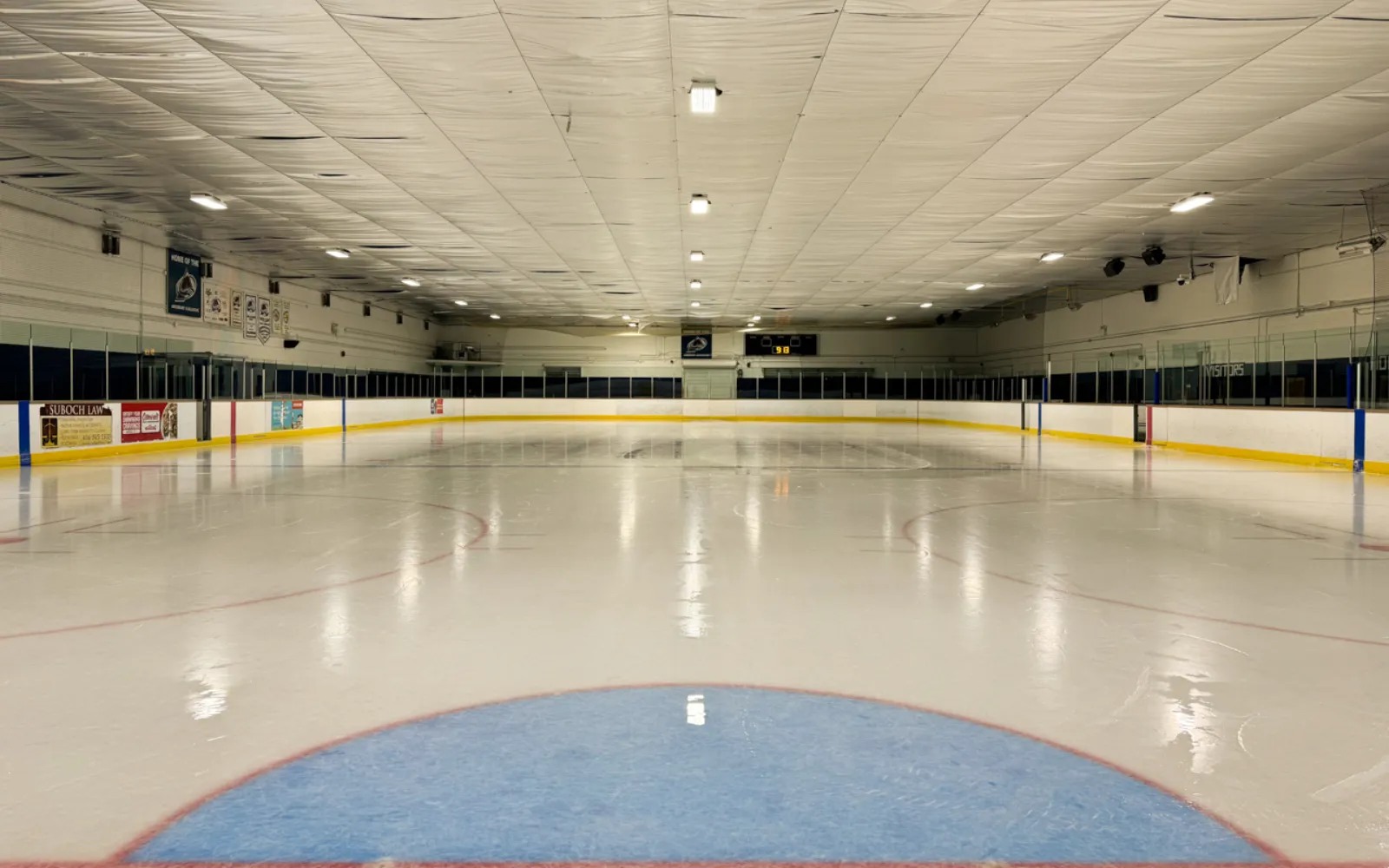 Full ice sheet with white vinyl ceiling above, red and yellow dasher-board padding, team/sponsor banner ('Home of the...') and scoreboard at the far end. — Amesbury Sports Complex