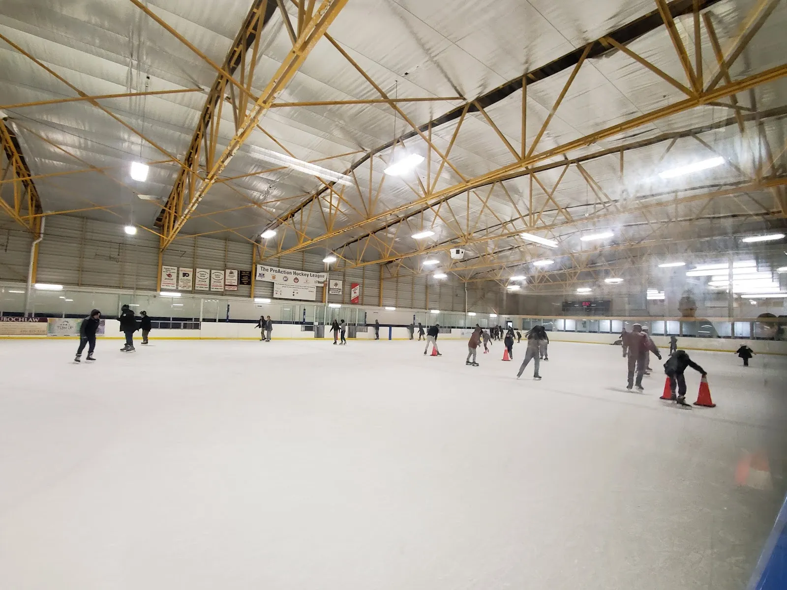 Wide interior view of the ice pad under yellow steel roof trusses, with public leisure skaters spread across the ice and orange traffic cones marking a lesson area. — Angela James Arena & Tennis Courts