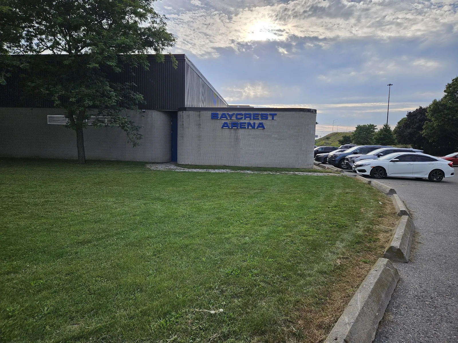 Side entrance of Baycrest Arena, a beige brick and dark metal-clad building with a blue "BAYCREST ARENA" sign above the door, parking lot alongside. — Baycrest Arena