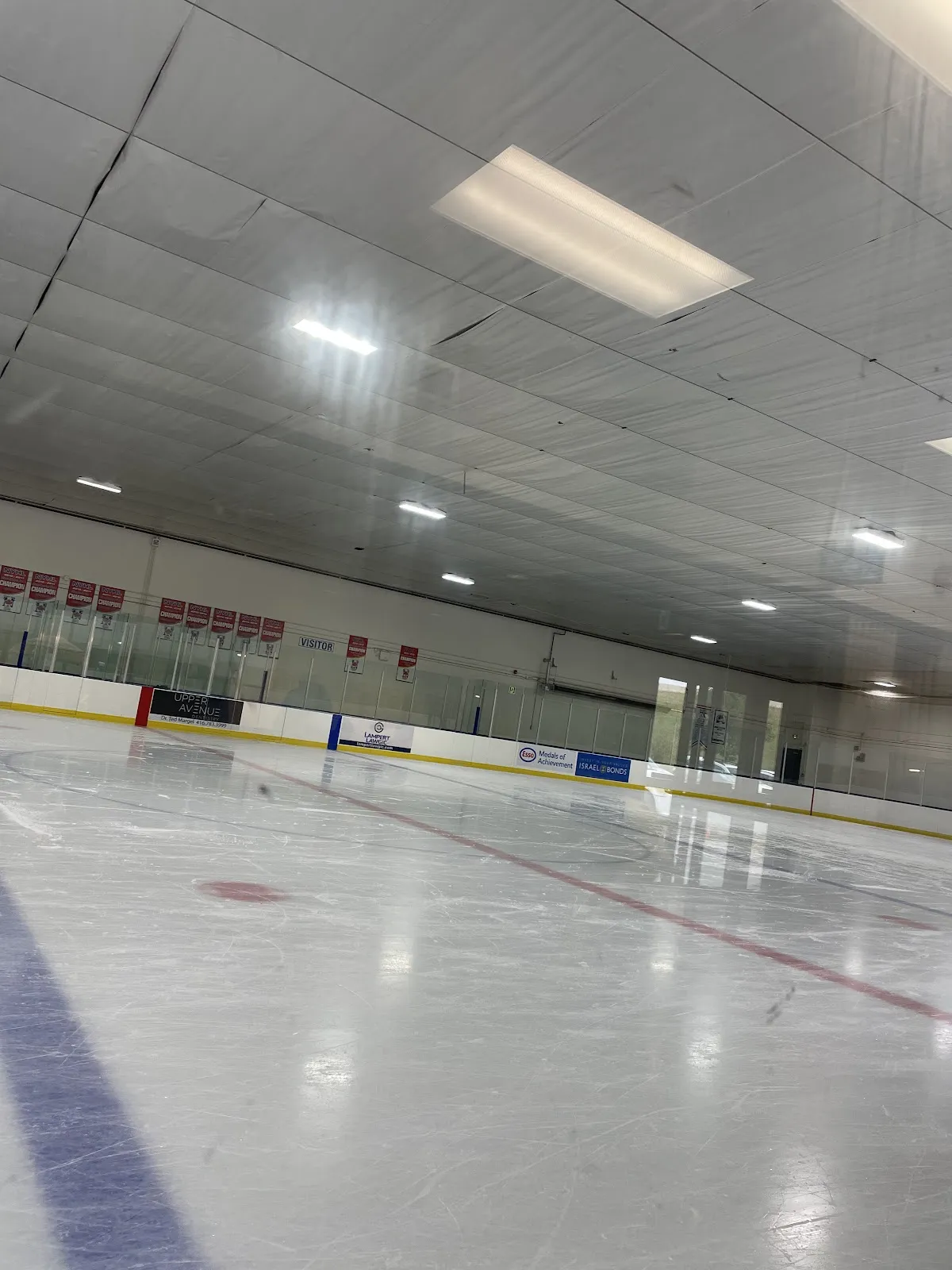 Empty ice pad viewed from ice level toward the boards, with sponsor/team championship banners along the wall and a "VISITOR" bench door; no people on the ice. — Baycrest Arena