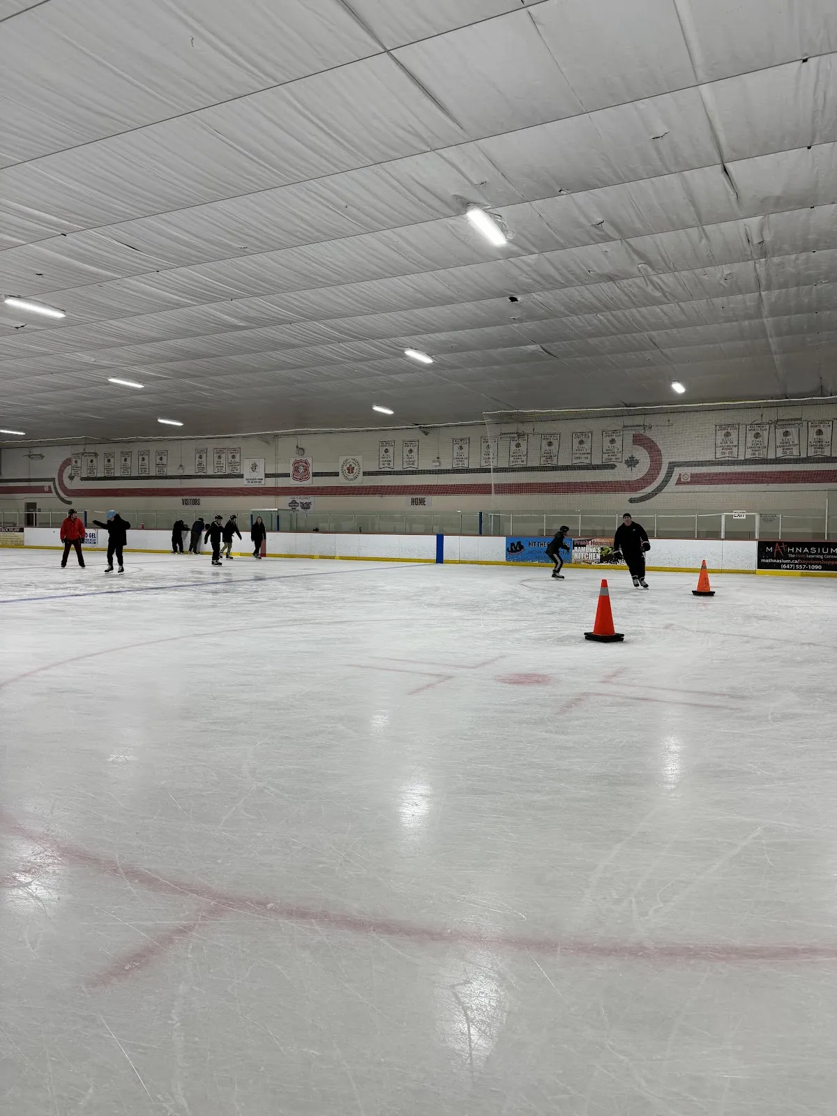 Wide view of the ice pad from ice level, with a handful of adult and youth leisure skaters and orange traffic cones near center ice; team/season championship banners line the back wall and a "bayviewshoppes" rink-board ad is visible. — Bayview Arena