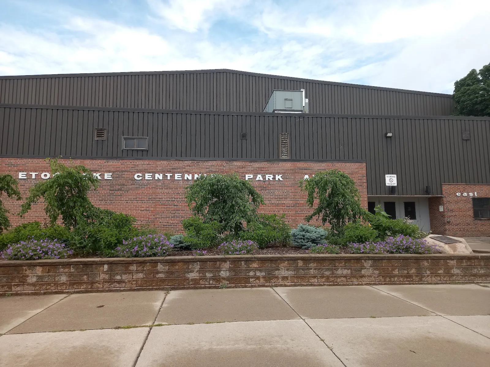 Brick and dark metal-clad facade reading "ETOBICOKE CENTENNIAL PARK A[RENA]" above a landscaped planting bed, with the east entrance doors visible at right. — Centennial Park Arena - Etobicoke