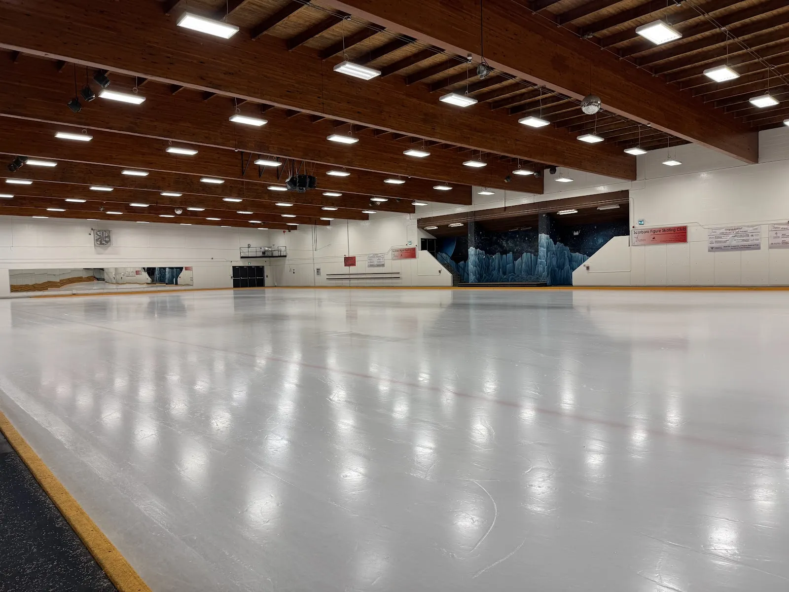 Pista techada vacía bajo un techo de vigas de madera, con un mural de una caverna de hielo en el muro del fondo y una manta del "Scarboro Figure Skating Club" en la pared lateral. — Centennial Recreation Centre - Scarborough