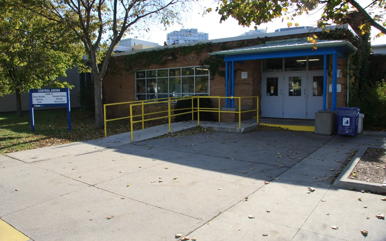 Front doors of Central Arena, a brick building with a blue canopy, a yellow-railed accessibility ramp and the arena sign on the lawn. — Central Arena