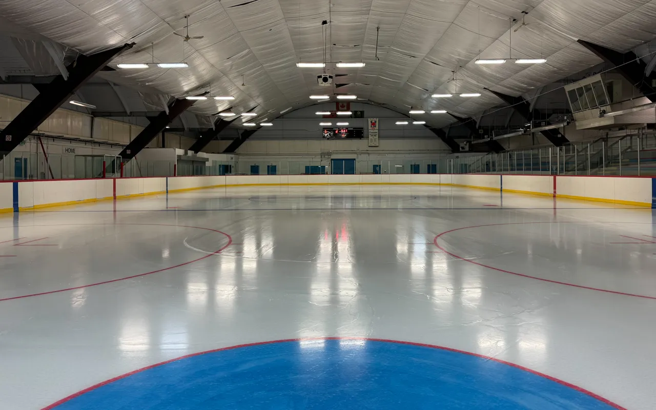An empty, freshly resurfaced ice sheet viewed from center ice toward the far boards, with the arched roof, rink lighting and a scoreboard visible, no people on the ice. — Chris Tonks Arena