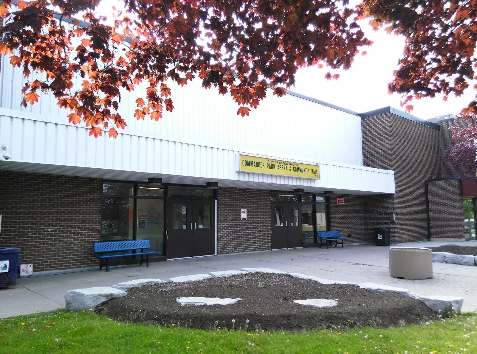 Entrance of the brick and metal-clad building under a yellow sign reading "CITY OF SCARBOROUGH COMMANDER PARK ARENA & COMMUNITY HALL 140", with benches by the doors. — Commander Recreation Centre