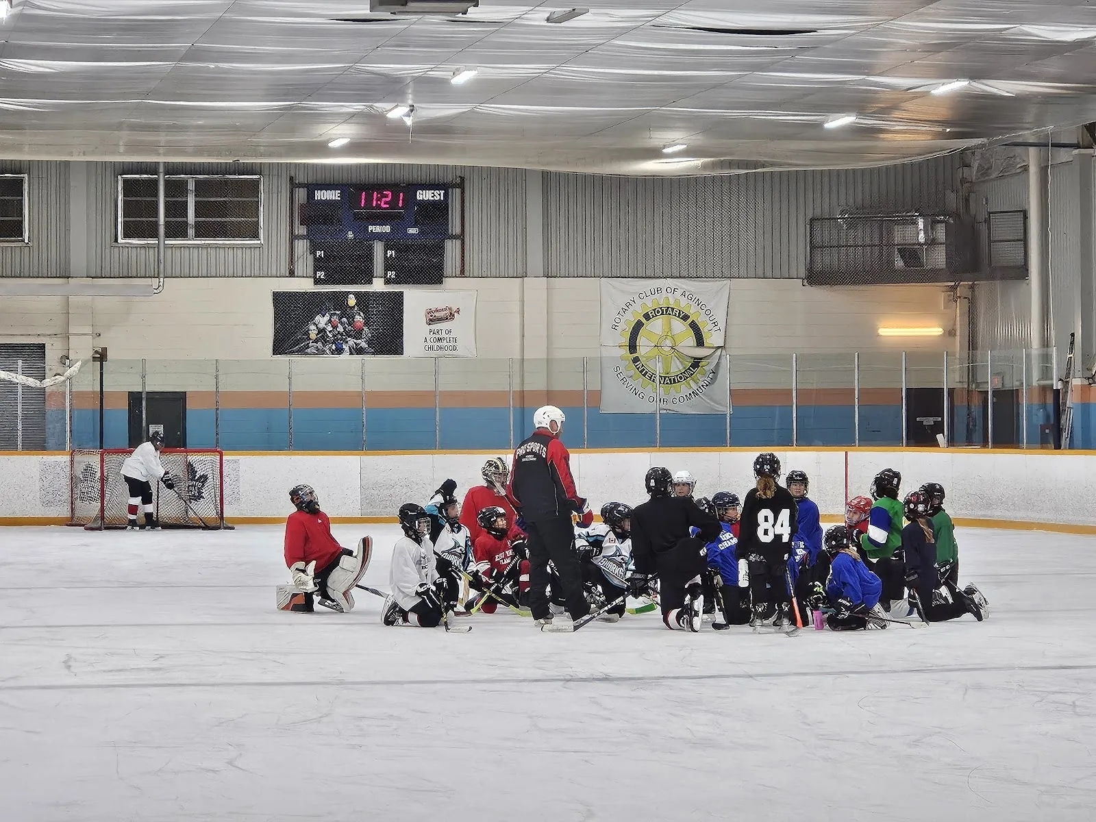 A practice on the sheet under a long ceiling with a scoreboard and club crest at the far wall, a group of children kneeling at one end. — Commander Recreation Centre