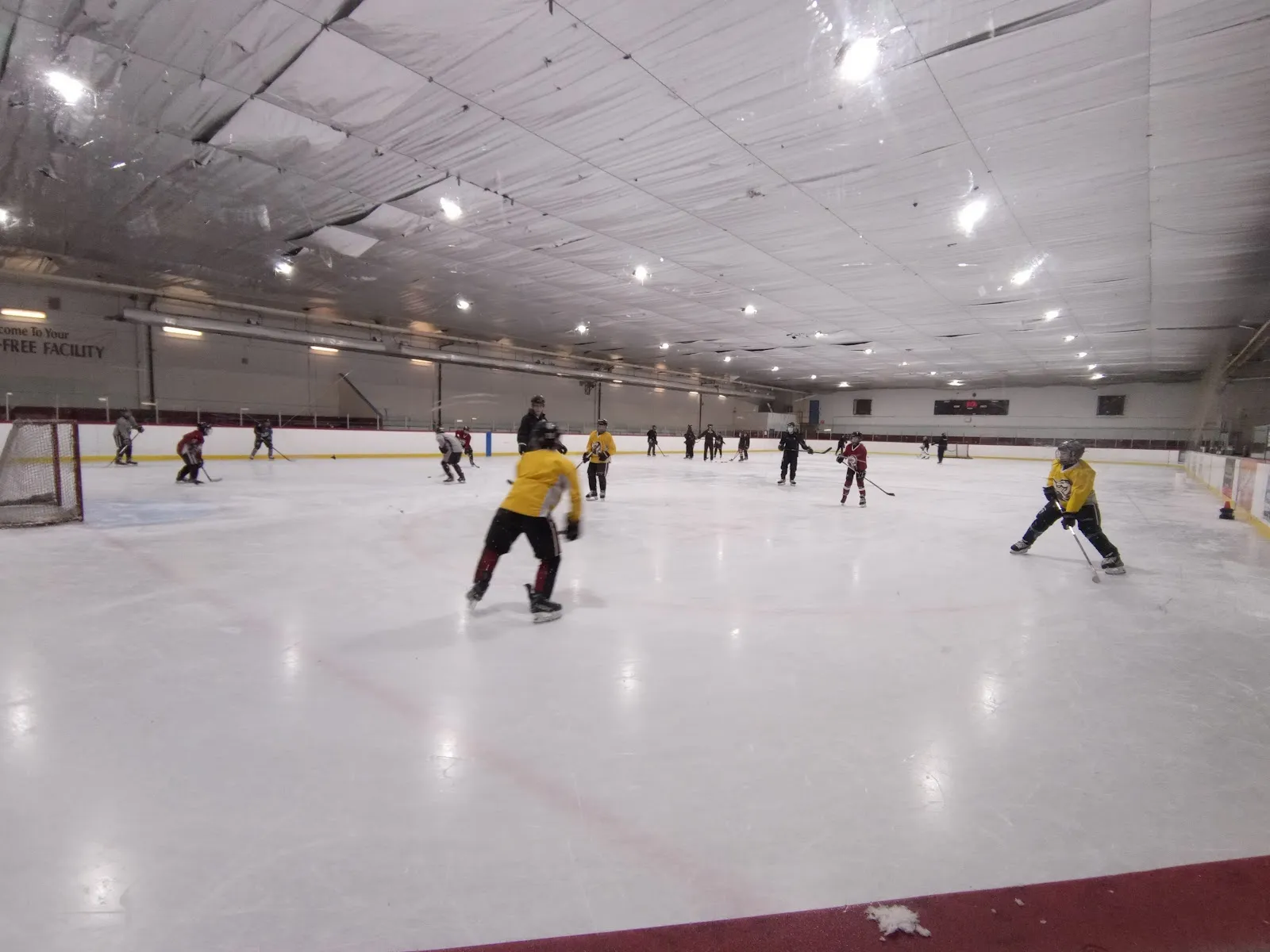 Un partido en una pista larga y plana visto desde las vallas, con jugadores repartidos por el hielo blanco y vallas con ribete rojo bajo un techo claro. — Don Mills Civitan Arena