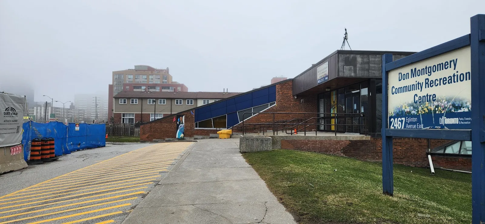 Andador principal hacia la entrada del Don Montgomery Community Recreation Centre, fachada de ladrillo con una nave en ángulo de azulejo oscuro y paneles azules sobre las puertas, el letrero de identificación del edificio en primer plano, tapiales de obra a la izquierda, sin personas. — Don Montgomery Community Recreation Centre