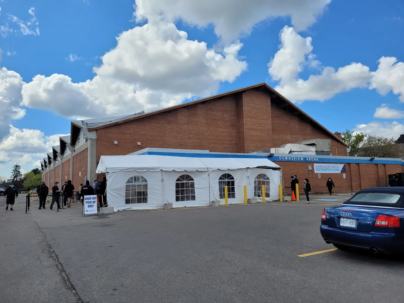 The brick Downsview Arena with a white arched entrance canopy at the front, a car park and a few people queuing under a blue sky. — Downsview Arena