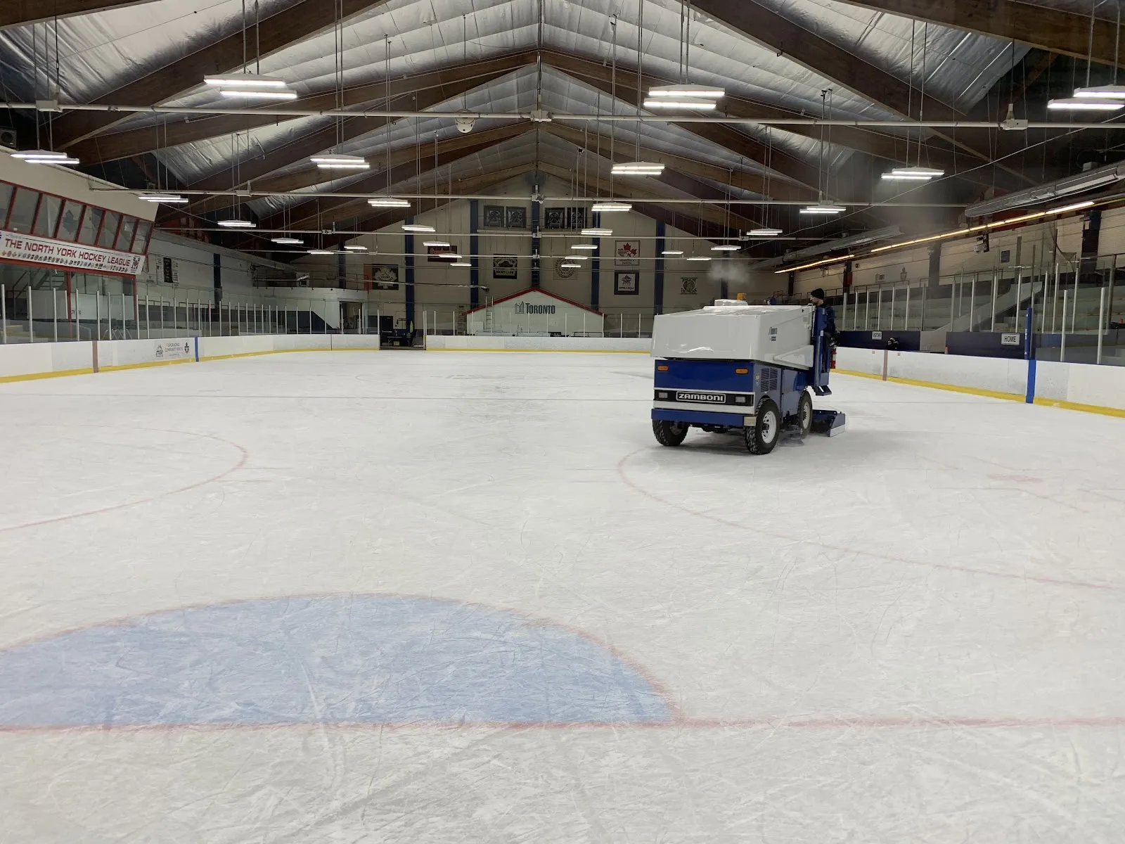 Full ice sheet under a peaked wood-truss ceiling, a Zamboni resurfacing the ice mid-rink; "THE NORTH YORK HOCKEY LEAGUE" banner and a Toronto crest banner hang on the end wall, with a "HOME" sign over the boards. — Downsview Arena