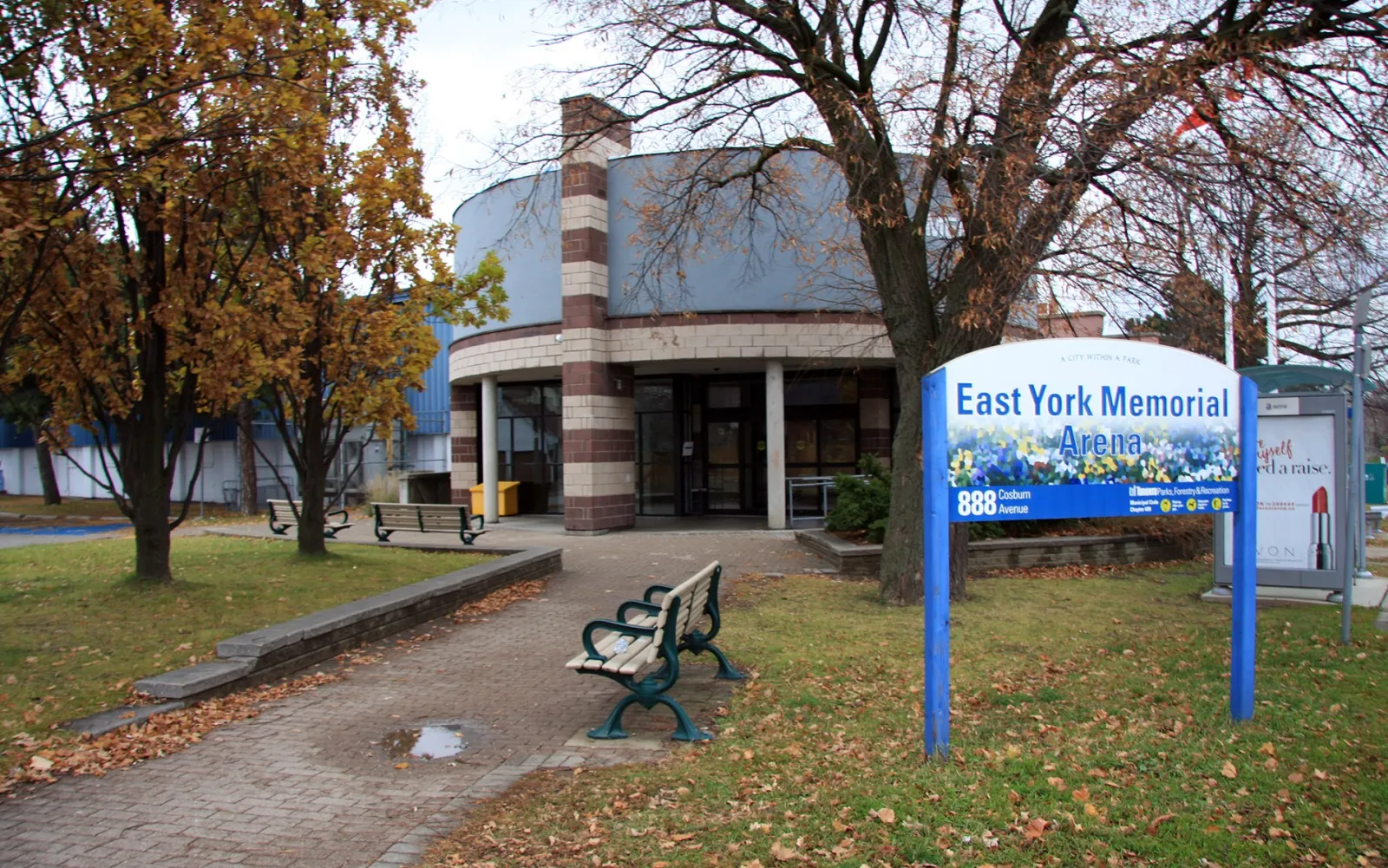 Entrada principal del East York Memorial Arena, un edificio de ladrillo con techo curvo visto desde el césped, con el cartel azul del arena delante. — East York Memorial Arena