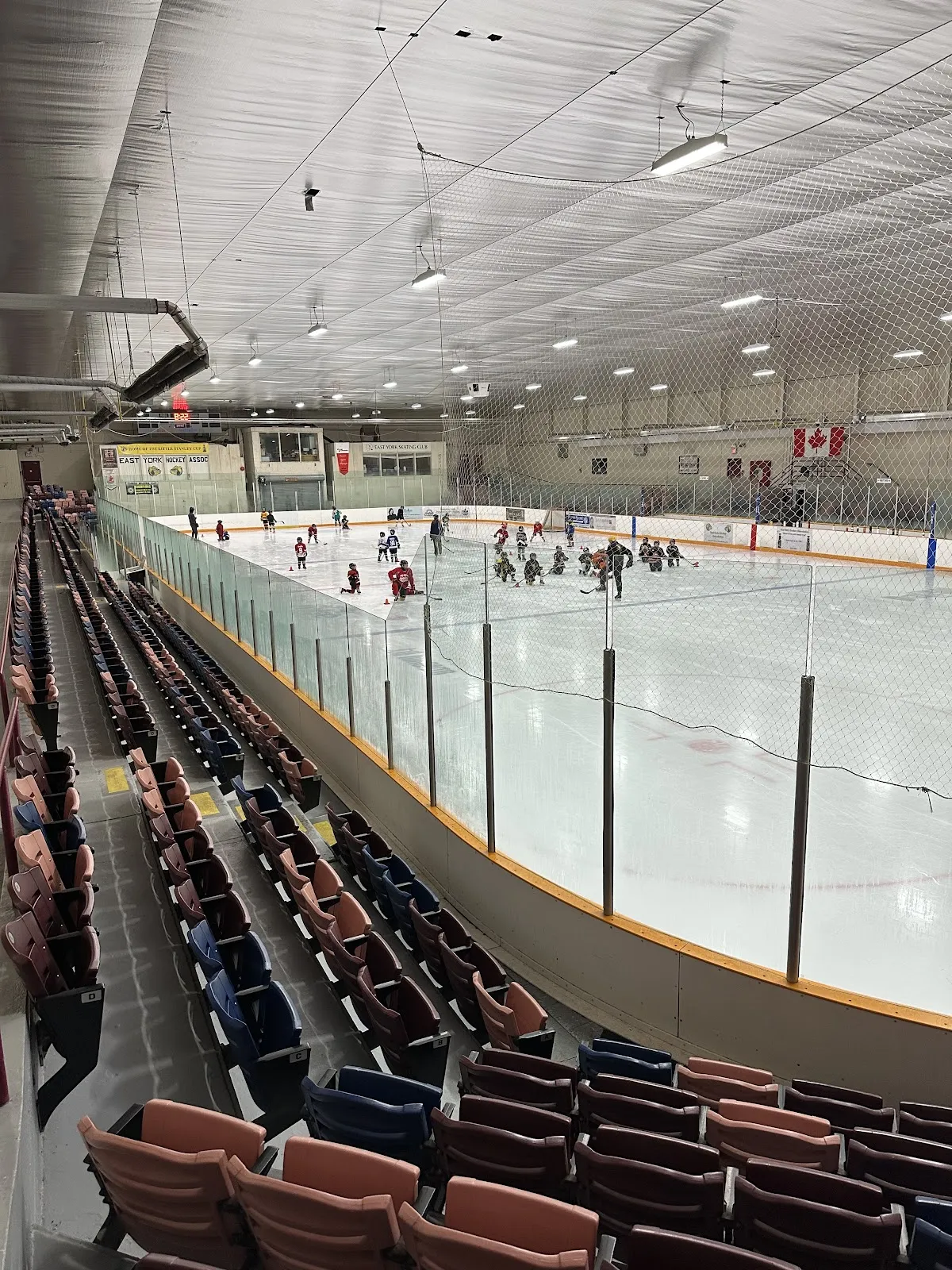 Toda la pista vista desde la tribuna elevada, con el cristal y las vallas en primer plano y una práctica de hockey juvenil en curso en la mitad lejana; mantas de "EAST YORK HOCKEY ASSOC" y "EAST YORK SKATING CLUB" y una bandera de Canadá cuelgan en el muro del fondo. — East York Memorial Arena