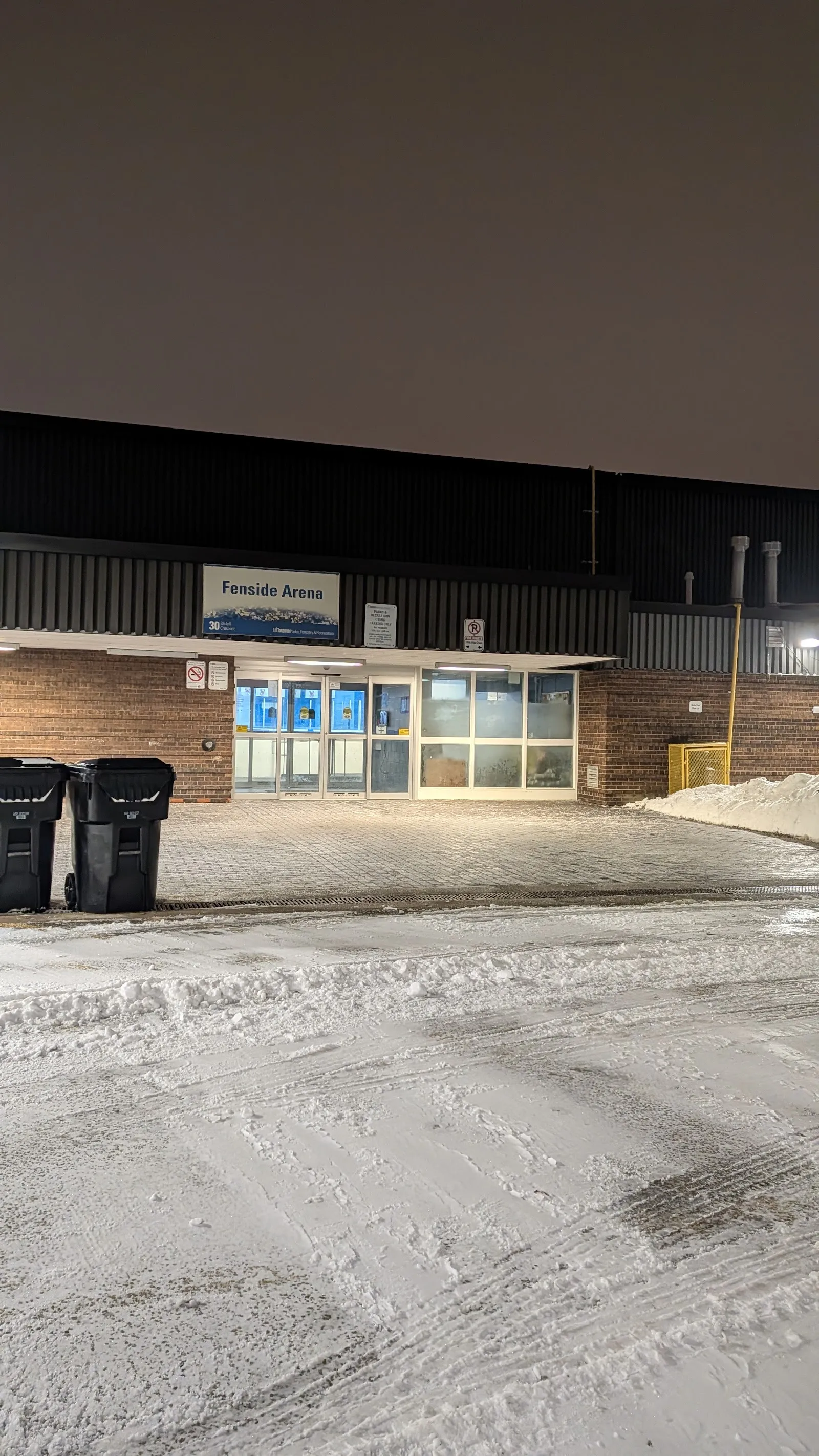 Night-time view of the Fenside Arena entrance, illuminated 'Fenside Arena' sign above brick facade and glass doors, freshly plowed snow in the lot, no people. — Fenside Arena