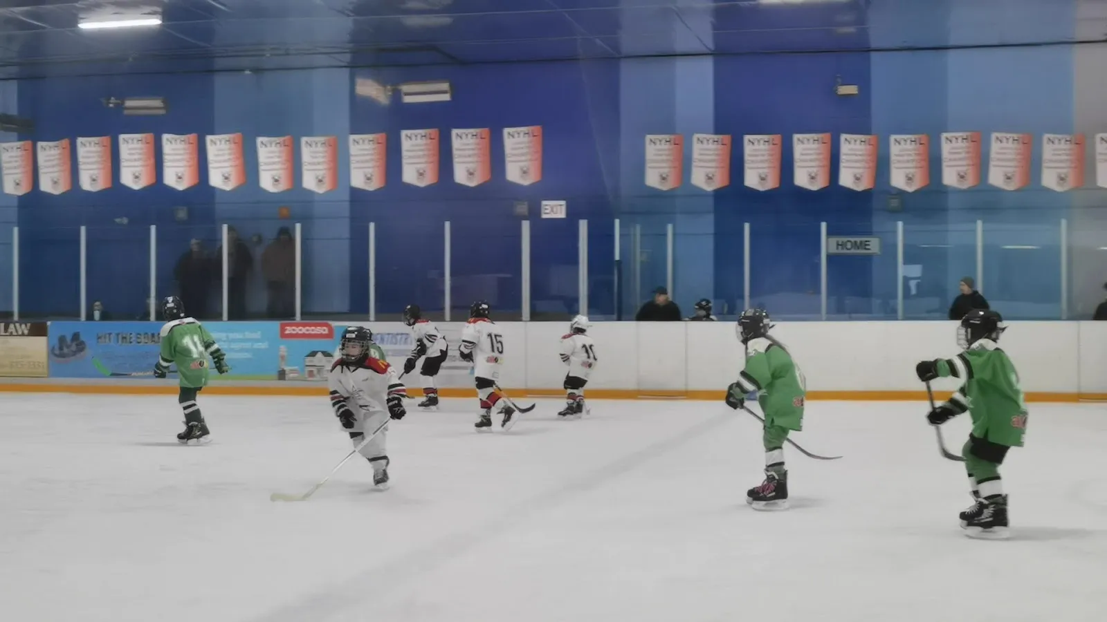 A game seen across the sheet at the boards, players spread near blue sponsor boards with a row of banners hung along the wall. — Fenside Arena