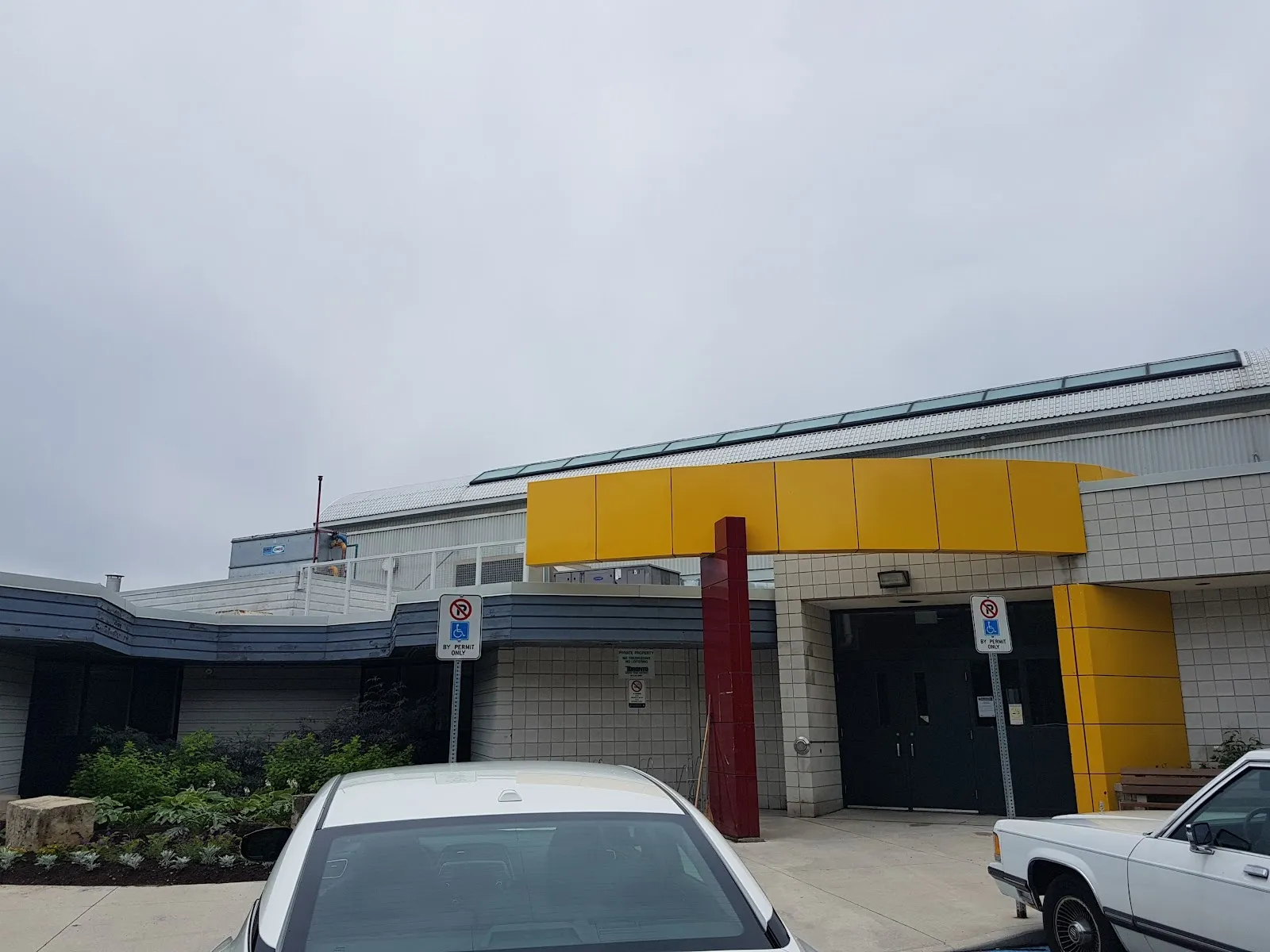 El centro comunitario gris con un toldo de entrada de marco amarillo y puertas oscuras, con autos estacionados al frente y una alberca al aire libre al lado. — Gord and Irene Risk Community Recreation Centre