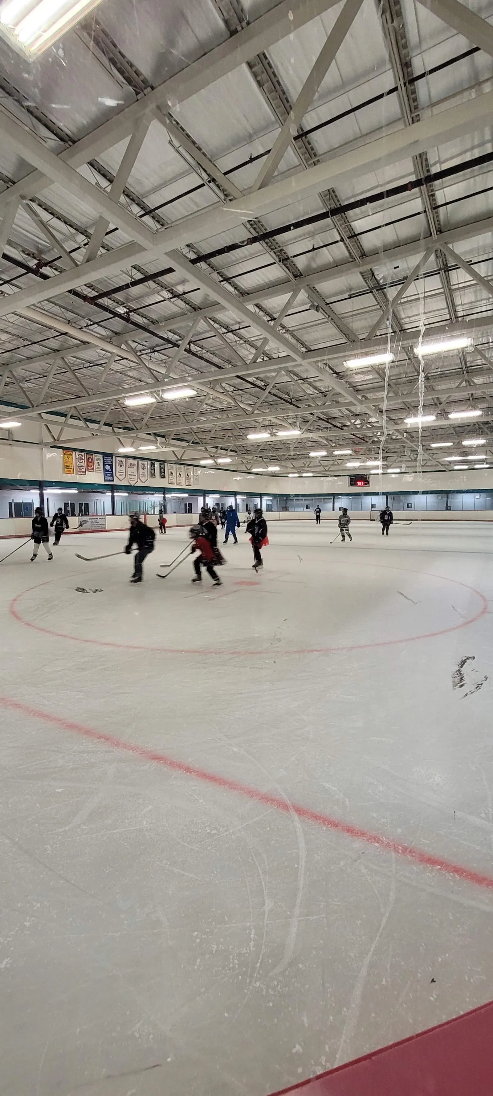Amplia vista interior de la pista de la arena Gord and Irene Risk durante una práctica de hockey, con las armaduras de acero del techo, mantas y la mayor parte de la pista. — Gord and Irene Risk Community Recreation Centre