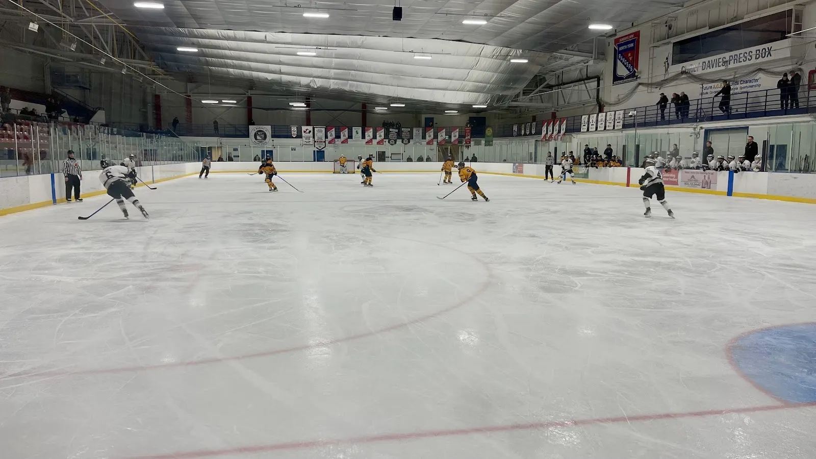 A wide flat view of the sheet from the end, players spread across the ice under a long steel-beam ceiling with banners along the far wall. — Herbert H. Carnegie Centennial Centre