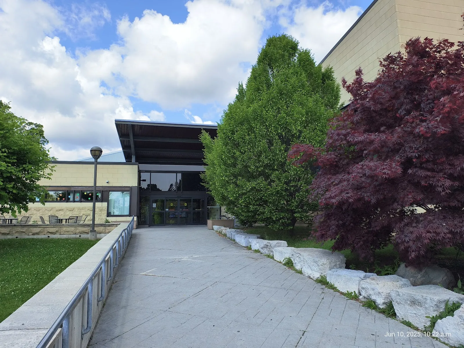 Entrada principal del Heron Park Community Centre, un edificio bajo de ladrillo color arena con un techo de marquesina oscuro sobre las puertas de vidrio de la entrada y un andador pavimentado. — Heron Park Community Recreation Centre